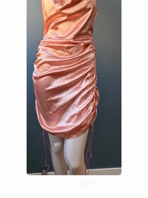 Champagnefärgad satin klänning - Snygg kort klänning i champagnefärgad satin med draperad framsida och tunna axelband. Klänningen har en omlottliknande effekt och en glansig finish som ger en lyxig känsla. Perfekt för dig som vill sticka ut.