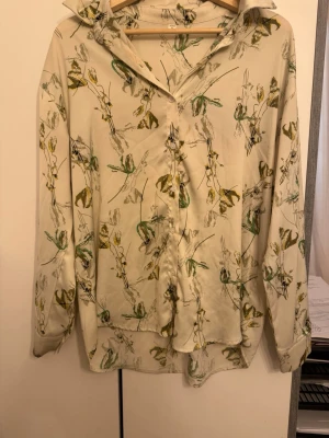 Beige blommig skjorta med grön detalj - Snygg beige skjorta med blommigt mönster i grönt och gult. Skjortan har klassisk krage, knäppning framtill och långa ärmar. Perfekt för dig som gillar en avslappnad men ändå stilren look.