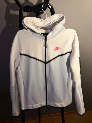 Vit Nike hoodie med dragkedja - Nike hoodie i vitt med svart dragkedja och svarta detaljer. Har en huva och rosa Nike-logga på bröstet. Tillverkad i mjukt och sportigt material, perfekt för en avslappnad och sportig stil.
