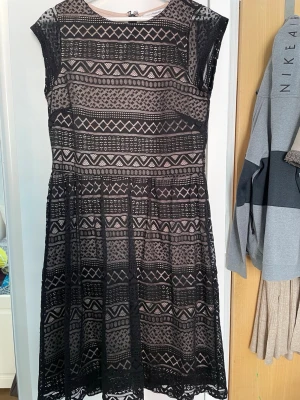 Svart spetsklänning med mönster - Säljer en svart spetsklänning med geometriskt mönster och beige underklänning. Klänningen har rund hals, är ärmlös och har en dragkedja i ryggen. Passformen är normal och längden går till knäna. Perfekt för dig som gillar unika detaljer och stilren design.