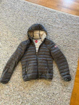 Mörkgrå dunjacka från Colmar - Snygg mörkgrå dunjacka från Colmar med huva och dragkedja framtill. Jackan har två fickor med dragkedja och Colmar-logga på ärmen. Insidan är ljusare grå och jackan är quiltad för extra värme. Perfekt för kyliga dagar.