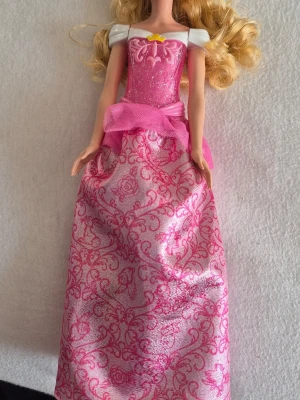 Barbiedocka i rosa prinsessklänning - Säljer en Barbiedocka med långt blont hår och en glittrig rosa prinsessklänning med vita puffärmar och dekorativt mönster. Klänningen har en rosa rosett i midjan och en liten gul detalj vid halsen. Perfekt för samling eller lek.