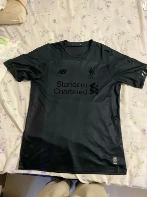 Svart Liverpool tröja från New Balance - Säljer en svart Liverpool FC t-shirt från New Balance med tryckt logga och sponsor på bröstet. Tröjan har rund hals och korta ärmar, tillverkad i ett lätt och sportigt material som passar perfekt till träning eller match.