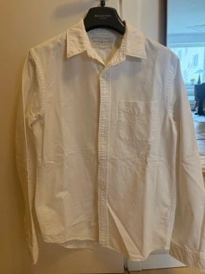 Vit skjorta från Ralph Lauren - Klassisk vit skjorta från Ralph Lauren i bomull med lång ärm, bröstficka och knappar framtill. Enkel och stilren design med normal passform och mjukt material. Perfekt till många olika looks.