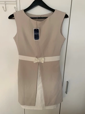 Beige kort klänning med rosett - Elegant beige kortklänning från Chopin med vit midjerem och söt rosettdetalj framtill. Klänningen är ärmlös och har en stilren, figurnära passform samt en slits framtill med vit underkjol. Perfekt för dig som gillar klassisk och stilren design.