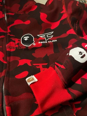 BAPE x FaZe Clan - Limited edition hoodie i röd och svart camo från samarbetet mellan BAPE och FaZe Clan. Stora loggor på bröstet och ryggen, dragkedja framtill och huva. Tillverkad i mjukt bomullsmaterial med ribbade muddar och detaljerade prints över hela plagget.