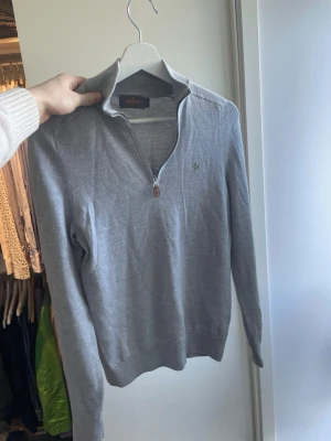 Grå zip-tröja från Morris - Snygg grå tröja från Morris med halvzip och hög krage. Tröjan har ett diskret broderat emblem på bröstet och ribbade muddar vid ärmslut och nederkant. Perfekt för lager-på-lager och stilrena outfits. Storlek S men har krympt så passar mer XS!