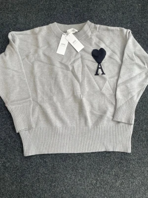 Grå stickad tröja från Ami - Snygg grå stickad tröja från Ami Alexandre Mattiussi med svart hjärta och A broderat på bröstet. Tröjan har rund hals, ribbade muddar och bred ribbkant nedtill. Perfekt för dig som gillar stilrena detaljer och mjuka material.