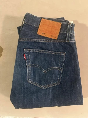 Levi's 501 mörkblå jeans - Klassiska Levi's 501 jeans i mörkblå denim med raka ben och knappgylf. Jeansen har fem fickor, kontrastsömmar och den ikoniska läderlappen bak i midjan. Perfekta för en tidlös och avslappnad stil.