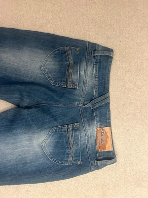 Blå jeans vintage - Snygga blå jeans från Dreamstar med slitningar och detaljerade sömmar på bakfickorna. Jeansen har dubbla knappar framtill och klassisk femficksdesign. Materialet är denim och färgen är en cool blå med ljusare tvättade partier.