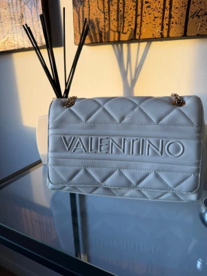 Väska från Valentino - Snygg vit handväska från Valentino med quiltat mönster och stora logotypen framtill. Väskan har guldfärgade kedjedetaljer och är tillverkad i ett slätt, mjukt material som ser ut som konstläder. Perfekt för dig som vill ha en trendig och stilren accessoar.