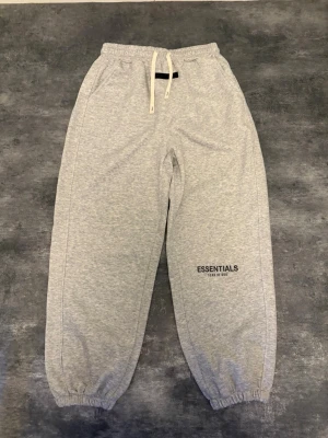 Grå Essentials mjukisbyxor - Säljer ett par grå mjukisbyxor från Fear of God Essentials med resår i midjan och benslut, samt vita dragsnören. Byxorna har sidofickor och logga tryckt på vänster ben. Perfekta för en chill och avslappnad stil.