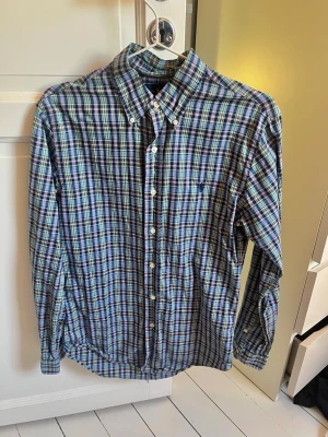 Rutig skjorta från Ralph Lauren - Klassisk rutig skjorta från Ralph Lauren i blå, grön och vit färgskala. Skjortan har lång ärm, knappar framtill och ikoniska broderade loggan på bröstet. Tillverkad i mjuk bomull och har en tidlös button-down krage.