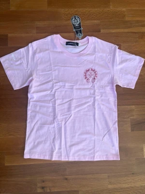 ChromeHearts T-shirt  - Ljusrosa Chrome Hearts T-shirt – storlek S Säljer en skön ljusrosa Chrome Hearts t-shirt. Har det stora cirkulära trycket på bröstet även där back med blomma och “Chrome Hearts” runt om, plus lite litet tryck på vänster ärm. T-shirten är mjuk och skön, klassisk passform Har hängmärket kvar, helt oanvänd helt ny. Storlek S. Skick: superbra, aldrig använd. Kan mötas upp eller skicka med frakt. Fråga på om du vill ha fler bilder