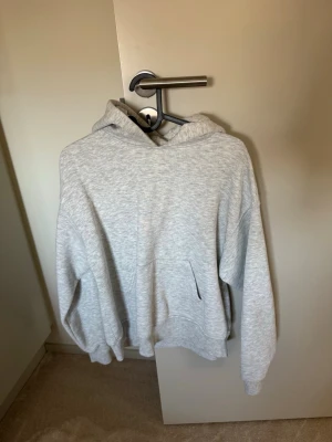 Ljusgrå hoodie med huva - En stilren ljusgrå hoodie med huva och magficka. Hoodien har en avslappnad passform, ribbade muddar vid ärmslut och nederkant. Perfekt för en chill och bekväm look. Materialet är mjukt och känns skönt mot huden.