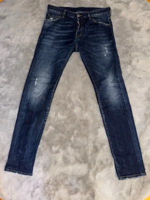 Mörkblå slim fit jeans från Dsquared2 - Snygga mörkblå slim fit jeans från Dsquared 2 med slitningar och tvättade detaljer. Klassisk femficksmodell med läderpatch bak i midjan. Jeansen har smal passform och är tillverkade i denim med lätt stretch för extra komfort.