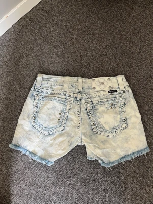 Ljusblå denimshorts med nitar - Säljer ett par ljusblå denimshorts från True Religion med slitna kanter och coola nitar längs fickorna. Shortsen har fem fickor, låg midja och en tvättad, nästan blekt look som ger en avslappnad vibe. Perfekta för sommaren!