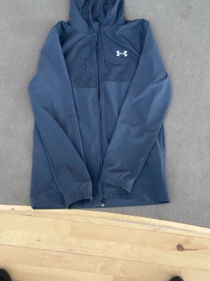 Mörkblå vindjacka från Under Armour - Snygg mörkblå vindjacka från Under Armour med huva och dragkedja framtill. Jackan har logga på bröstet och stor text på ryggen. Tillverkad i lätt och vindtätt material, perfekt för blåsiga dagar. Elastiska muddar vid ärmsluten och praktiska fickor.