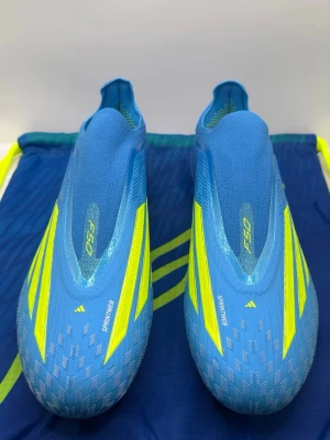 Blåa Adidas F50 fotbollsskor - Snygga blåa Adidas F50 fotbollsskor med gula detaljer och ikoniska stripes. Skorna har en strömlinjeformad design utan snörning och är tillverkade i lätt syntetmaterial för maximal fart på planen. Perfekta för dig som vill sticka ut och prestera på topp.