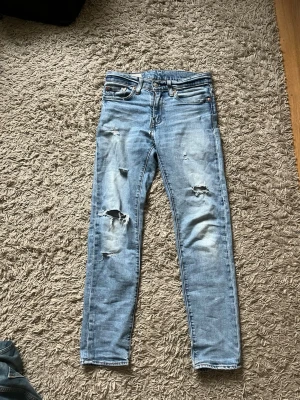 Ljus blå levis jeans - Säljer ett par ljusblå jeans från Levi's med slitningar och hål på benen för en trendig look. Klassisk femficksmodell med läderpatch bak i midjan och dragkedjegylf. Perfekta för dig som gillar en avslappnad och cool stil.                 Storlek: w28 L30.  