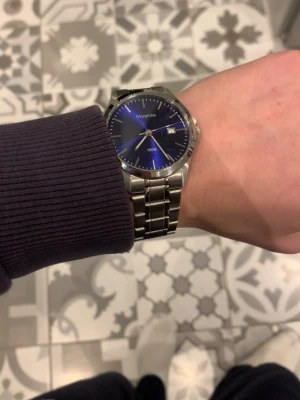 Montine klocka med blå urtavla - Stilren klocka från Montine med metallarmband i silver och en snygg blå urtavla. Klockan har datumvisning och klassisk rund form. Perfekt accessoar för dig som gillar en tidlös look.