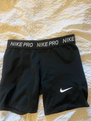 Svarta Nike Pro träningsshorts - Säljer ett par svarta Nike Pro träningsshorts med elastisk midja där det står 'NIKE PRO' i vitt. Shortsen har en tajt passform och är tillverkade i ett stretchigt funktionsmaterial som passar perfekt för gym eller löpning. Klassisk vit Nike-logga på benet.