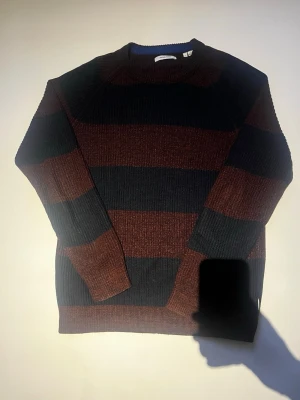 Randig stickad tröja Jack & Jones - Mysig stickad tröja från Jack & Jones i storlek L. Tröjan har breda horisontella ränder i mörkbrunt och svart, rund halsringning med blå insida och ribbstickade muddar. 