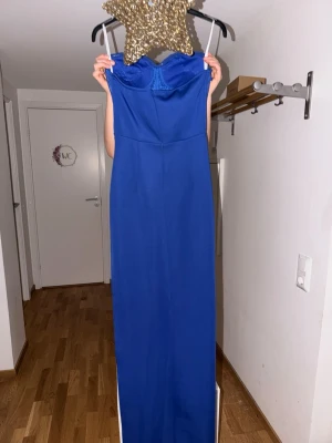 Blå axelbandslös långklänning - Snygg blå axelbandslös långklänning med glittrande stenar och pärlor upptill. Klänningen har en figurnära passform och är tillverkad i ett glansigt material som ger en elegant look. Perfekt för speciella tillfällen.