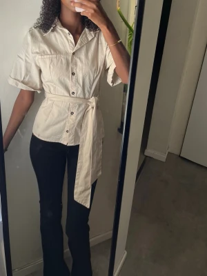 Beige skjorta med knytskärp - Stilren beige skjorta med korta ärmar, knappar framtill och ett brett knytskärp i midjan. Skjortan har klassisk krage och bröstficka, perfekt för en trendig och avslappnad look. från zara helt ny i storlek xs🌼