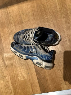 Nike Air Max Plus blå/vit sneakers - Nike Air Max Plus sneakers i blått och vitt med ikoniska vågmönster på ovandelen. Skorna har vita snören, svart sula med synliga Air-enheter och en liten nike logga på sidan. Materialet är mesh och syntet, perfekt för dig som gillar streetwear och sportig stil. Båda skorna har små hål på sidorna som finns med på bilden. Skorna är köpt för 2500 priset är ej satt 100%