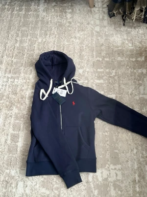Marinblå hoodie från Polo Ralph Lauren - Snygg marinblå hoodie från Polo Ralph Lauren med dragkedja, vit dragsko och klassisk röd logga på bröstet. Tillverkad i mjuk bomullsblandning och har känguruficka framtill. Perfekt för en avslappnad och stilren look.