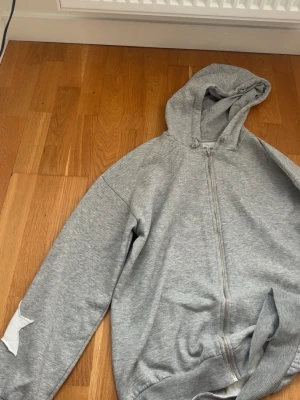 Grå hoodie med vita stjärnor - Säljer en grå zip-hoodie med huva och vita stjärnpatchar på ryggen och ena ärmen. Hoodien har dragkedja framtill och ribbade muddar vid ärmslut och nederkant. Perfekt för en chill och avslappnad stil. Materialet känns mjukt och bekvämt.