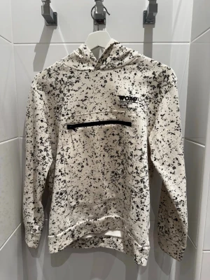 Vit hoodie med svart splattermönster från Woxo - Säljer en vit hoodie från Woxo med svart splattermönster och en stor ficka med dragkedja framtill. Hoodien har tryckt logga på bröstet och är tillverkad i mjukt bomullsmaterial. Perfekt för dig som gillar streetwear och unik design.