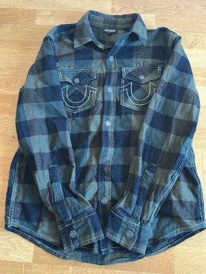 Rutig skjorta från True Religion - Snygg rutig skjorta från True Religion i blått och grönt med stora bröstfickor och vit kontrastsöm. Skjortan har lång ärm, knappar framtill och en stor logga med tryck på ryggen. Perfekt för en avslappnad streetstil.