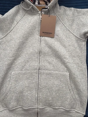 Grå hoodie med dragkedja Burberry - Grå hoodie från Burberry med dragkedja, fickor fram och klassisk huva med Burberrys ikoniska rutmönster på insidan. Tillverkad i mjuk bomullsblandning och har diskret broderad logga på bröstet. Perfekt för en clean och stilren look.