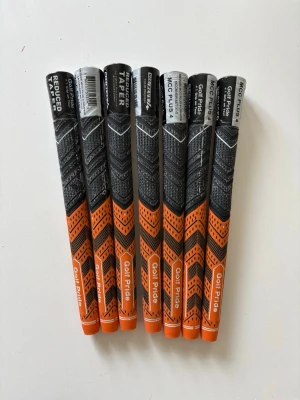 Golf Pride grepp 7-pack - Säljer sju stycken Golf Pride grepp i svart och orange. Greppen har texturerat mönster för extra grepp och komfort, samt märkning 'Reduced Taper'. Perfekt för dig som vill uppgradera dina golfklubbor med snygga och funktionella grepp.