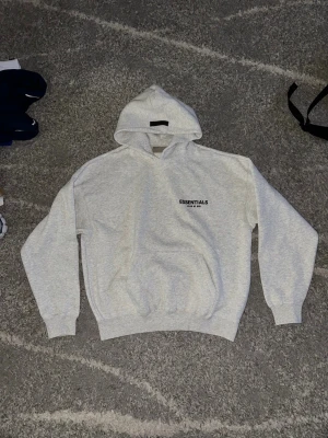Grå Essentials hoodie Fear of God - Säljer en ljusgrå hoodie från Essentials Fear of God med svart logotyp på bröstet och stor text på ryggen. Hoodien har huva, ribbade muddar och en klassisk känguruficka. Perfekt för dig som gillar streetwear och vill ha något stilrent. Storlek: det står att det är storlek S men passformen är för M eller L och det är anledningen till att jag säljer den, det skulle kunna passa på S men isåfall får man förvänta sig att den sitter lite baggy