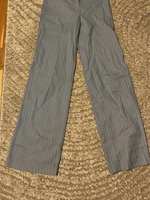 Ljusblå randiga kostymbyxor Monki - Snygga ljusblå kostymbyxor från Monki med tunna svarta vertikala ränder. Byxorna har rak passform och klassisk midja, perfekta för en stilren look. Tillverkade i ett mjukt tyg som känns bekvämt hela dagen.