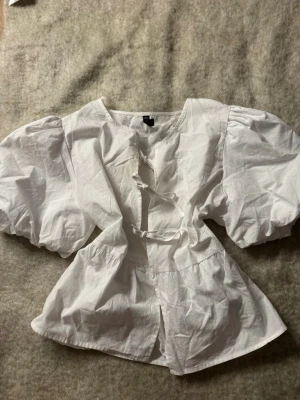 Vit blus med puffärmar och knytning - Superfin vit blus med stora puffärmar och knytning framtill. Blusen har en lös passform och är tillverkad i ett lätt bomullstyg som känns luftigt. Perfekt för dig som gillar en romantisk och trendig stil.