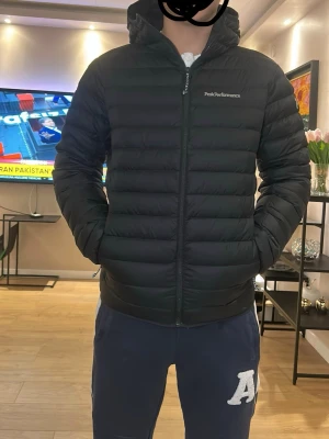 Svart dunjacka från Peak Performance - Snygg svart dunjacka från Peak Performance med huva och diskret logga på bröstet. Jackan har quiltad design med horisontella sömmar, dragkedja framtill och två sidofickor. Perfekt för kalla vinterdagar och riktigt stilren look. Jag har bara inte etiketten på (har använt bara 2 gånger)
