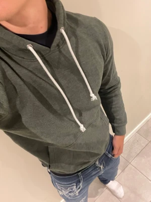 Mörkgrön hoodie med vita snören - Säljer en stilren mörkgrön hoodie med huva och vita dragsnören. Hoodien har en klassisk känguruficka framtill och ribbade muddar vid ärmslut och nederkant. Perfekt för en avslappnad look och passar till jeans eller joggers.💋Modellen bär storlek S och är 181 cm lång och väger 76 kg kom gärna vid frågor eller funderingar💋