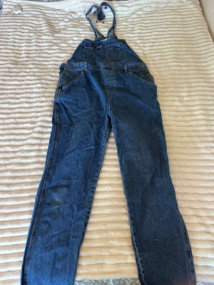 Blå denim hängselbyxa jumpsuit - Säljer en klassisk blå denim jumpsuit med hängslen och raka ben. Modellen har stora fickor framtill, bröstficka och två bakfickor. Tillverkad i slitstarkt bomullsjeansmaterial och har justerbara axelband för perfekt passform. Perfekt för en avslappnad och trendig look.