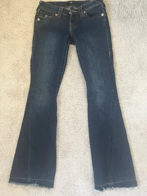 True Religion bootcut jeans blå - W23, Säljer ett par blå bootcut jeans från True Religion med klassiska kontrastsömmar och snygga fickdetaljer bak. Jeansen har låg midja, fem fickor och rå kant nedtill. Perfekta för dig som gillar en avslappnad men trendig look. Skriv innan ni köper dom är också sydda så dom passar nån runt 158