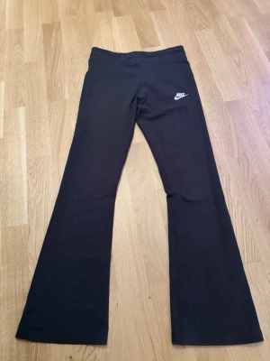 Svarta utsvängda leggings från Nike - Svarta leggings från Nike med utsvängda ben och klassisk vit logga på höften. Storleken är 156-166 den är stretchiga fast längden är lite kort. Själv är jag 166 och skulle säga att den är kort för mig. Skulle säga att den passar tjejer med längden 142-158. Har används bara 1 gång.
