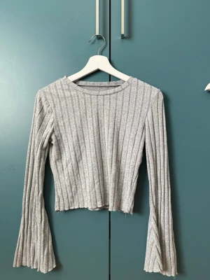 Tröja  - Säljer en grå ribbad topp med lång ärm och utsvängda ärmslut. Toppen har en croppad passform och ett stilrent randigt mönster. Perfekt för dig som gillar en enkel men trendig look.
