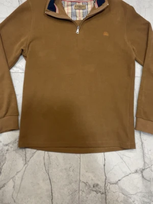 Brun halvzip från Burberry - Snygg brun långärmad tröja från Burberry med halv dragkedja och klassiskt rutigt foder i kragen. Tröjan har broderad Burberry-logga på bröstet och ribbade muddar vid ärmslut. Perfekt för en stilren och avslappnad look.