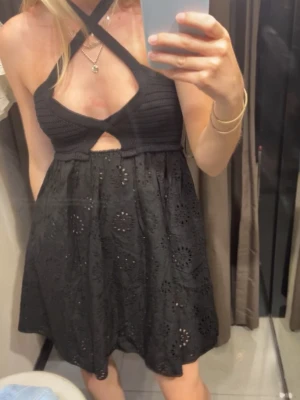 Svart kort klänning med broderi - Snygg svart kort klänning med broderade detaljer och utstansat mönster. Klänningen har omlott halterneck och en lätt utställd kjol. Perfekt för dig som vill ha en trendig och unik look.