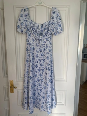 Vit blommig långklänning med puffärm - Superfin långklänning i vit med blått blommönster. Klänningen har puffärmar, knytning vid bysten och en slits framtill. Perfekt för sommarens alla tillfällen och ger en romantisk vibe. Materialet känns lätt och luftigt.