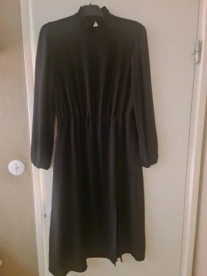 Svart långärmad klänning med slits - Stilren svart klänning med lång ärm och hög hals. Klänningen har markerad midja och en diskret slits framtill för extra rörelsefrihet. Perfekt för dig som gillar en enkel men elegant look.