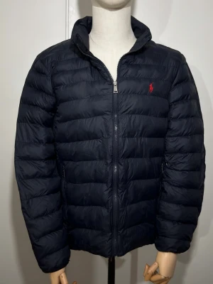 Mörkblå pufferjacka från Polo Ralph Lauren - Snygg mörkblå pufferjacka från Polo Ralph Lauren med klassisk röd logga på bröstet. Jackan har dragkedja framtill, ståkrage och två sidofickor. Tillverkad i lätt och vadderat material som håller dig varm under kalla dagar.